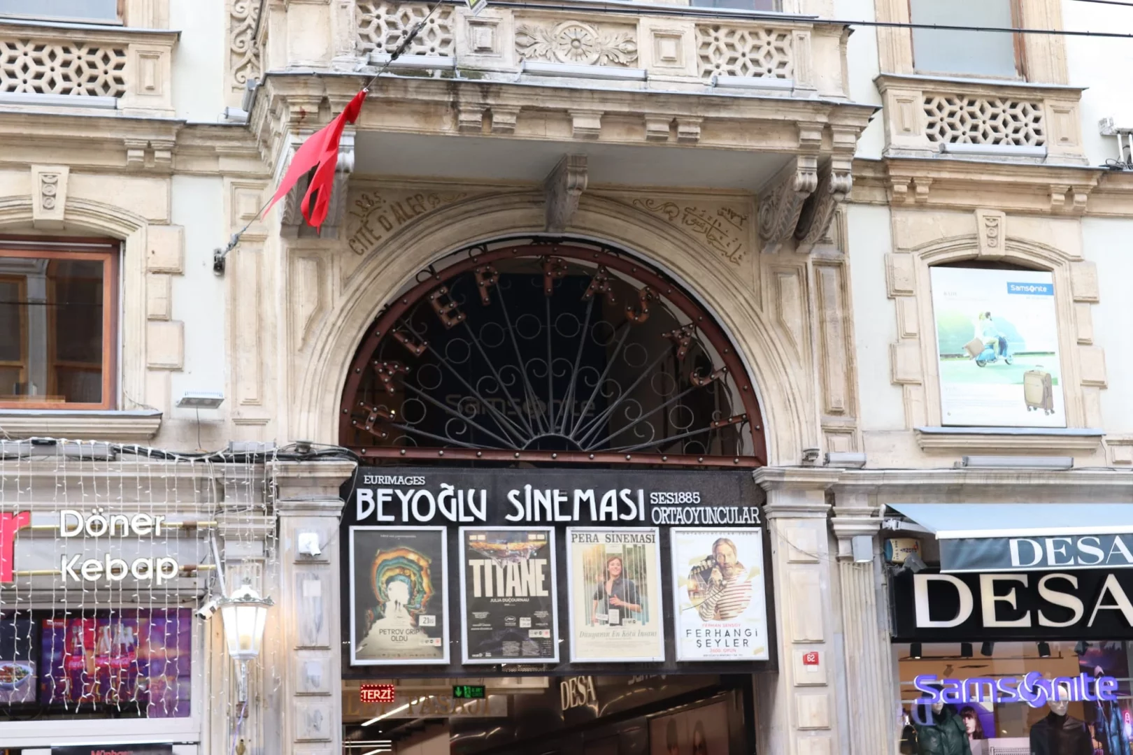 İstiklal Caddesi üzerindeki tarihi bir binanın görkemli cephesini yakından gösteren bu görselde, kemerli bir girişin üzerinde Grand Pera yazan zarif demir işçiliği, kırmızı bir bez parçasının asılı olduğu üst kat balkon süslemeleri, alt kısımda ise belirgin bir şekilde Beyoğlu Sineması tabelası ile Titane ve Ferhangi Şeyler gibi filmlerin afişleri dikkat çekiyor.