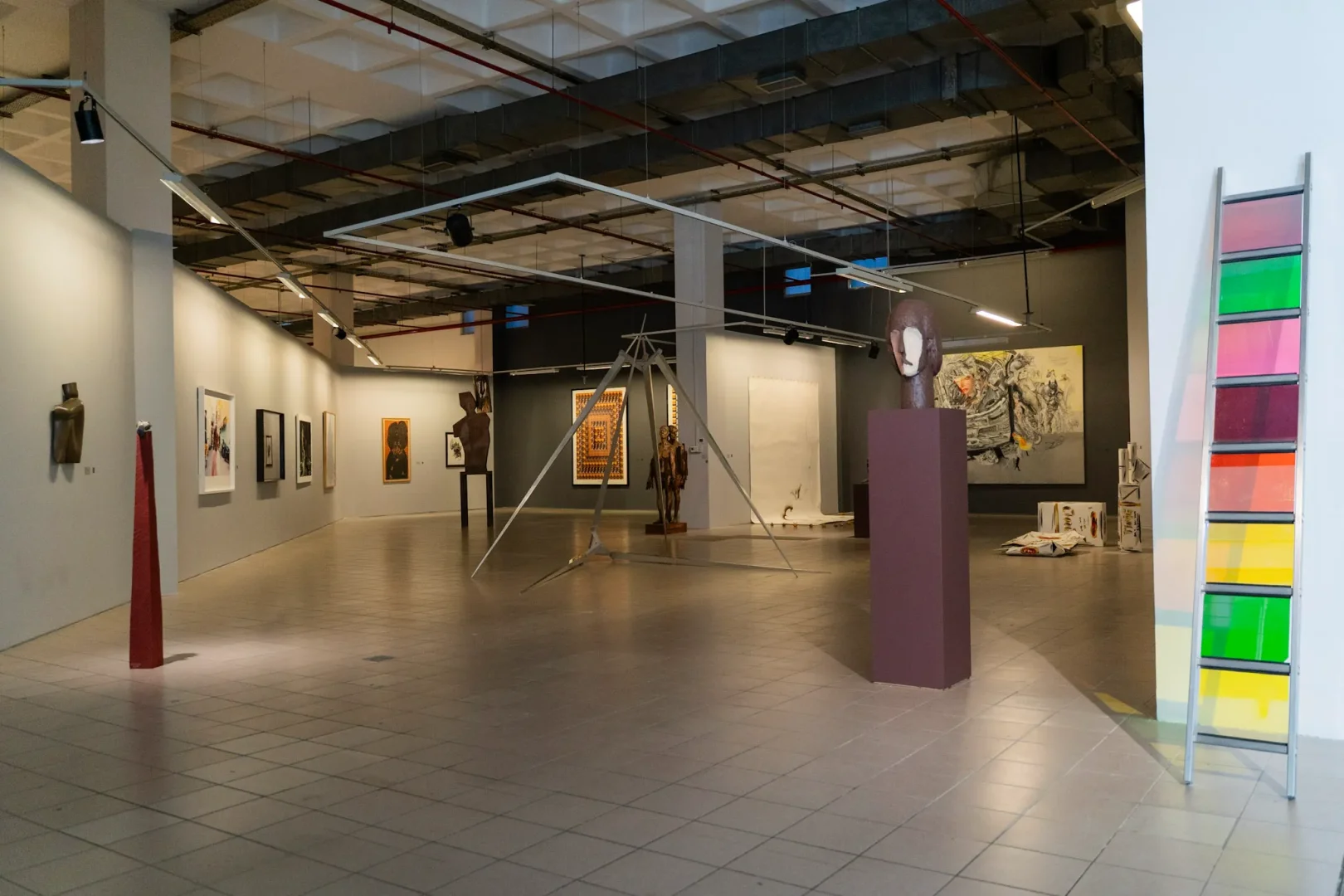 Elgiz Müzesi'ndeki modern sanat sergisinden, heykellerin ve tabloların sergilendiği geniş, aydınlık salonda, sağda pembe, yeşil, turuncu gibi şeffaf panellerden oluşan merdiven şeklinde dikkat çekici bir enstalasyon görülüyor.