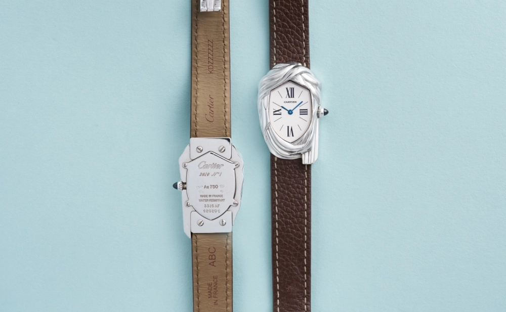 Açık mavi bir zeminde sergilenen, Sotheby's müzayedesindeki rekor satışla ilgili Cartier Cheich kol saatlerinden ikisi: Soldaki saatin ışık yansıtan gümüş renkli kasa arkasında Cartier yazısı ve Au 750 gibi üretim detayları belirginleşirken, sağdaki kol saati, ikonik türban benzeri gümüş kasası, beyaz oval kadranı, mavi ibreleri ve koyu kahverengi deri kayışıyla önden tüm ihtişamıyla gözüküyor.