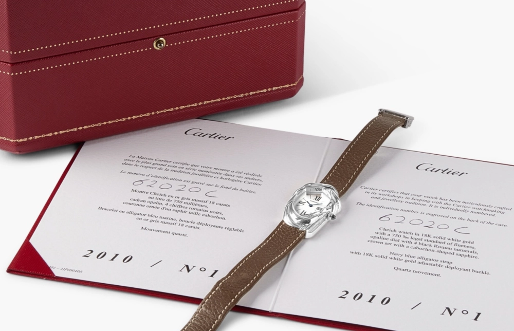 Sotheby's müzayedesinde rekor kıran ve 18 ayar beyaz altından üretilmiş, erimiş görünümlü eşsiz kasasıyla dikkat çeken Cartier Cheich kol saati, kahverengi deri kayışıyla, üzerinde 62020C kimlik numarası yazılı beyaz bir sertifikanın üzerinde ve kırmızı kutusunun önünde duruyor.