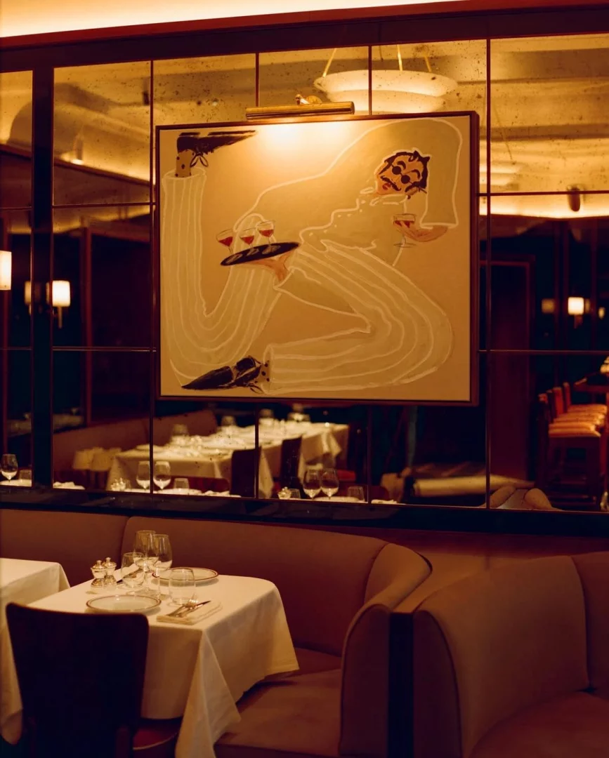 Loş ışıklı, şık bir restoran iç mekanında, bıyıklı bir garson figürünü ve kadehli bir tepsiyi betimleyen büyük, stilize bir tablo, aynalı bir duvarda asılı dururken, ön planda beyaz örtülü, zarif sofralar ve bej döşemeli koltuklar mekana sıcak bir atmosfer katıyor.