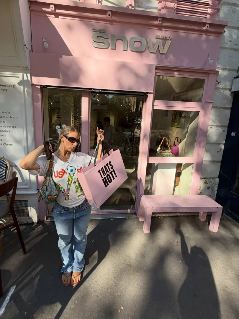 Pembe cepheli Chez Snow Bunny mağazasının önünde poz veren genç bir kadın, Thats Hot! yazan pembe bir alışveriş çantasını ve küçük bir omuz çantasını tutarken, beyaz tişörtü, kot pantolonu ve terlikleriyle güneşli bir günde şık bir görünüm sergiliyor.