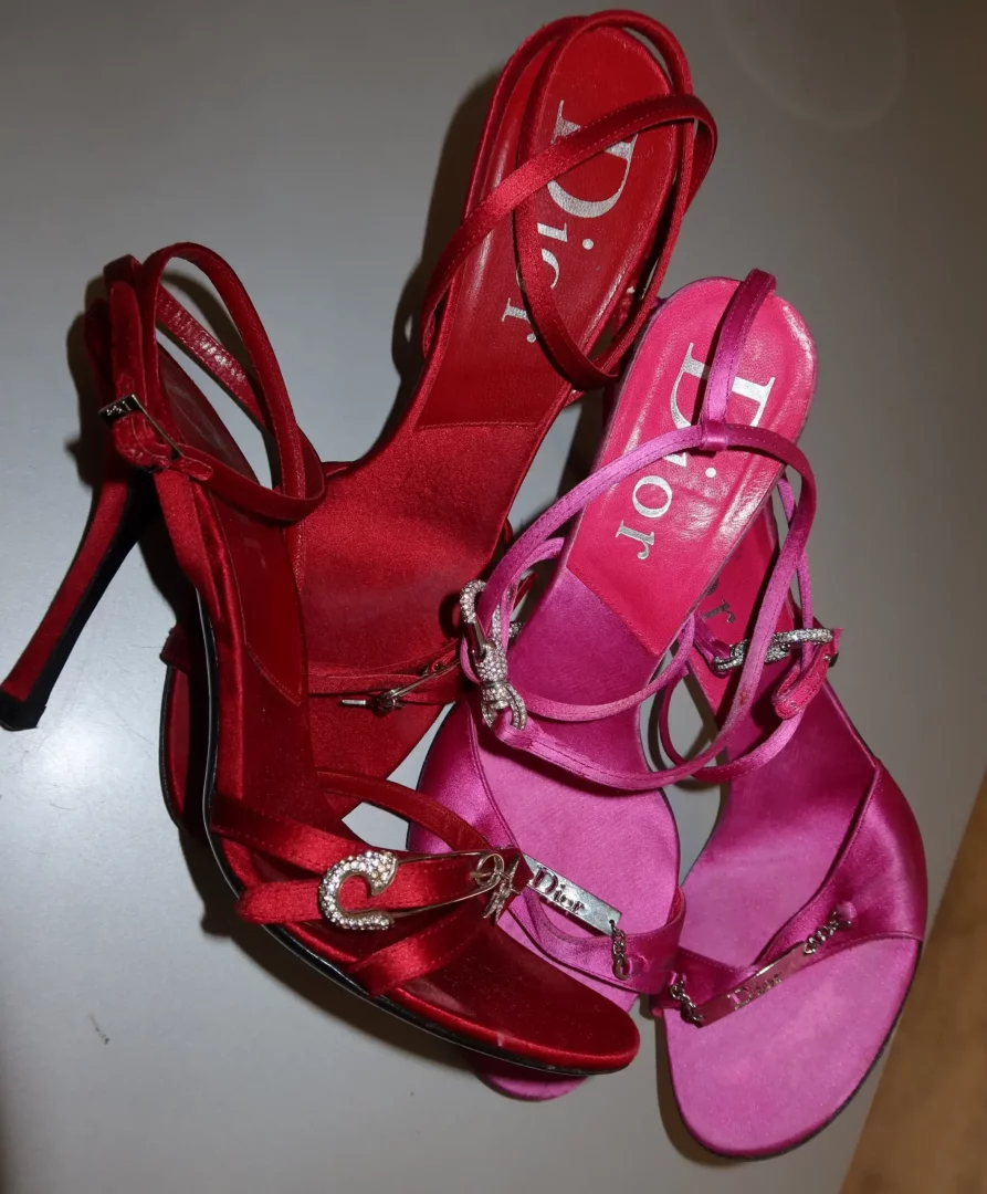 Parlak saten kumaştan yapılmış, biri kırmızı yüksek topuklu, diğeri fuşya renkli ince bantlı lüks Dior sandaletler, her ikisinin de iç tabanında Dior logosu belirgin bir şekilde görülürken, kırmızı olanı ışıltılı bir emniyet iğnesi detayıyla, fuşya olanı ise metal bir Dior charm ile süslenmiş olarak gri bir zeminde yan yana duruyor.