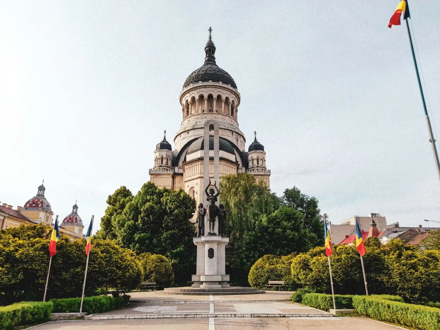 Cluj-Napoca şehrinde yer alan ve büyük bir kubbesi ile dikkat çeken ihtişamlı bir katedralin önünde, etrafı yemyeşil ağaçlar ve çalılarla çevrili, heykelleri olan anıtsal bir yapı ile direklerde dalgalanan Romanya bayrakları huzurlu bir şehir manzarasını oluşturuyor.
