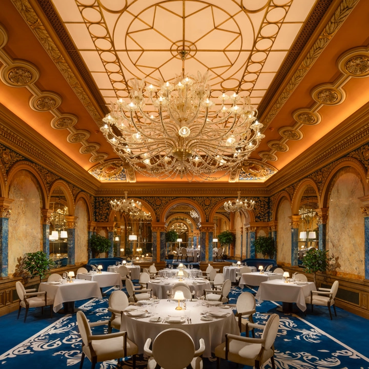 Corinthia Bükreş'in lüks restoranında, altın rengi varak süslemeli yüksek tavanı, tavandan sarkan devasa kristal avizeleri, beyaz örtülü şık yuvarlak masaları ve mavi desenli halısıyla göz alıcı bir yemek salonu yer alıyor.
