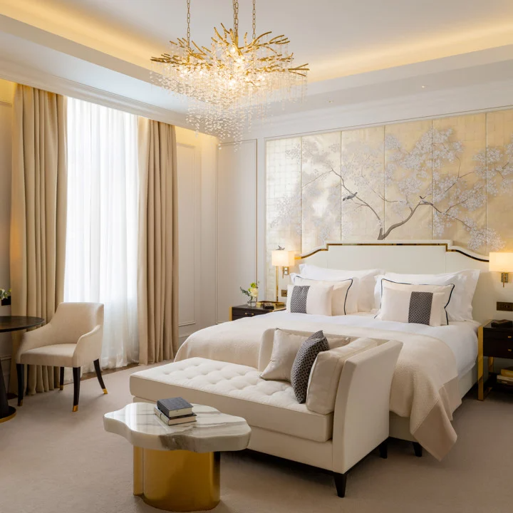Corinthia Bucharest'in krem ve altın tonlarındaki zarif bir lüks yatak odasında, duvarı süsleyen kuş desenli ağaç motifli paneller, başucundaki büyük yatak, yatak önünde şık bir bank ve tavandan sarkan ışıltılı bir avize modern konforu vurguluyor.