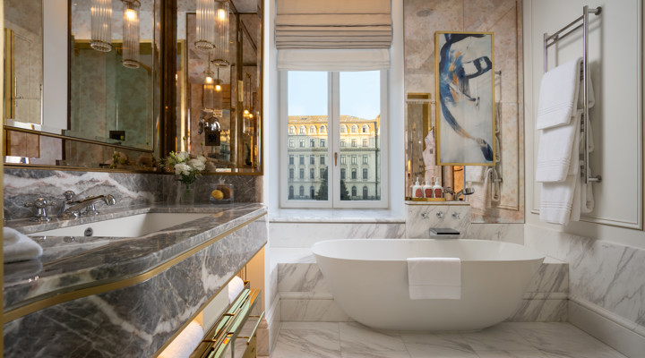 Corinthia Bucharest otelinin lüks banyosunda, şık gri mermer tezgahlı bir lavabo, pencereden görünen zarif tarihi bina manzarası sunan bağımsız bir küvet ve duvarda modern bir tablo ile beyaz havluların asılı olduğu bir havluluk göze çarpmaktadır.
