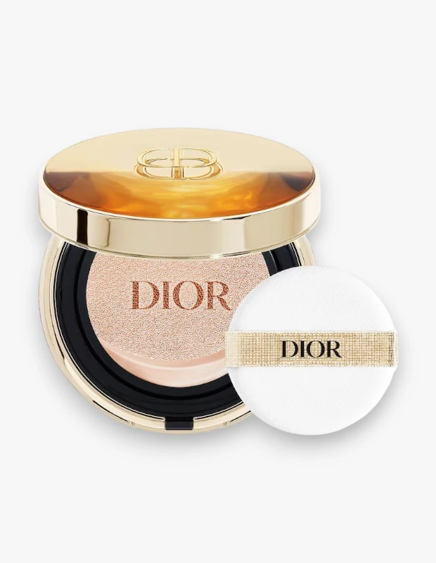 Parlak altın rengi kapağında kabartma logo, içinde ise DIOR yazılı açık bej rengi fondötenin görüldüğü şık bir Dior Prestige Le Cushion Teint de Rose fondöten kutusu ve hemen yanında DIOR yazısıyla süslenmiş beyaz makyaj süngeri bulunuyor.