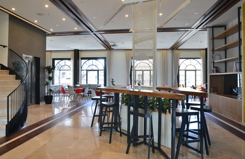 Divan Brasserie Beyoğlu'nun aydınlık ve modern iç tasarımında, ahşap şeritli parlak mermer zemin, kavisli merdiven, asansör, yeşil bitkilerle ayrılmış yüksek ahşap bar masaları ve çeşitli renklerde sandalyelerin olduğu geniş oturma alanları, ayrıca kemerli pencereler ve ahşap tavan kirişleri dikkat çekiyor.