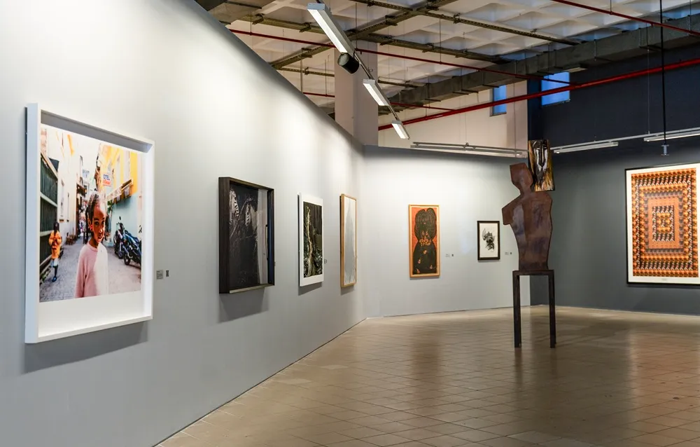Elgiz Müzesi'ndeki güncel bir sergiden etkileyici bir kare; açık gri duvarlarda farklı boyutlarda tablolar ve bir fotoğraf sergilenirken, zemin üzerinde yükselen metal bir heykel, modern sanat eserlerinin buluştuğu bu geniş ve aydınlık galerinin atmosferini yansıtıyor.