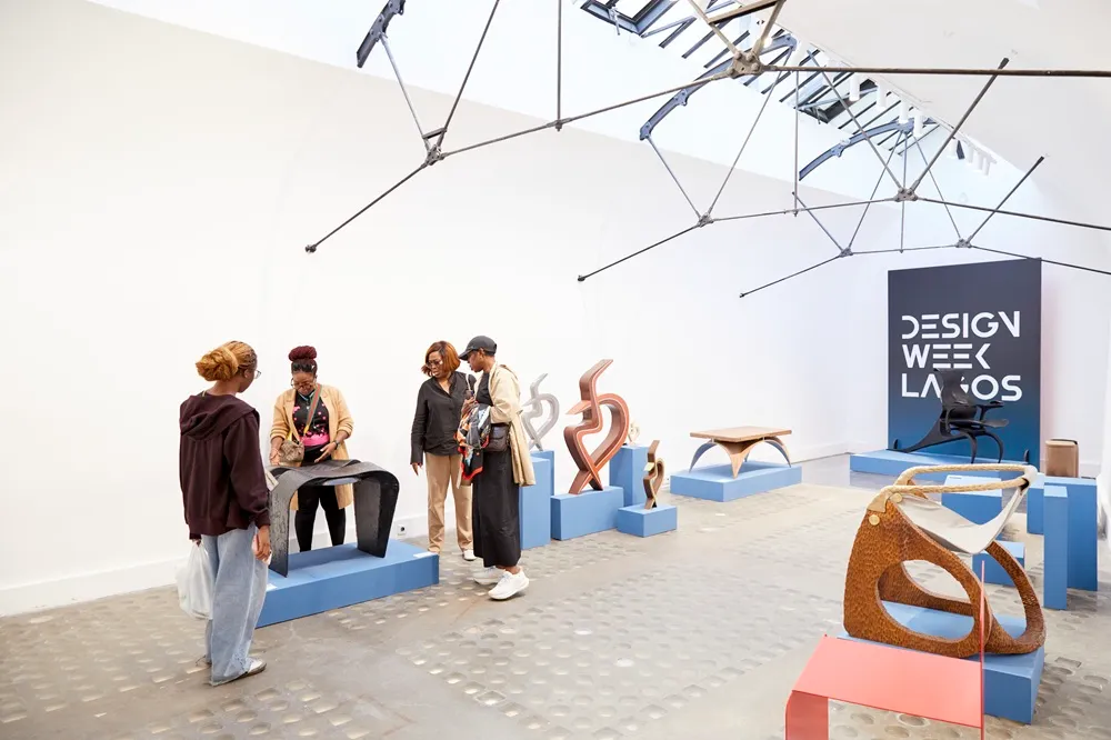 Geniş ve aydınlık bir galeride, Lagos Tasarım Haftası sergisi kapsamında sunulan, farklı tasarımlara sahip modern mobilya parçaları ve soyut heykelsi objeler mavi podyumlar üzerinde sergilenirken, ziyaretçiler bu özgün ve yenilikçi eserleri dikkatle inceliyorlar.