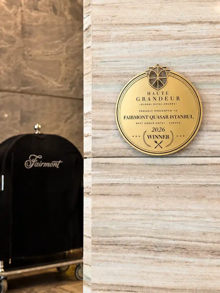 Fairmont Quasar Istanbul otelinin, açık renk mermer duvarda asılı duran altın renkli Haute Grandeur Global Hotel Awards plaketinde 2026 yılı Avrupa'nın En İyi Şehir Oteli ödülünü gururla kazandığı belirtilirken, sol tarafta markanın amblemini taşıyan siyah bir bagaj arabası kısmen görülüyor.
