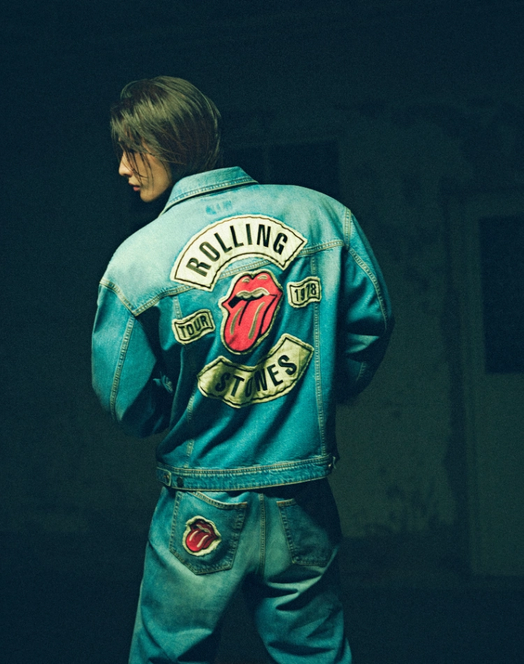 Rolling Stones'un ikonik dil logosu ve 'Tour 1978' yazılı yamalarıyla süslenmiş, arkası dönük bir kişinin giydiği mavi denim ceket ve uyumlu kot pantolon, markaların göze çarpan yeni tasarımlarını ve rock 'n' roll ruhunu yansıtan işbirliklerini modern bir estetikle sunuyor.