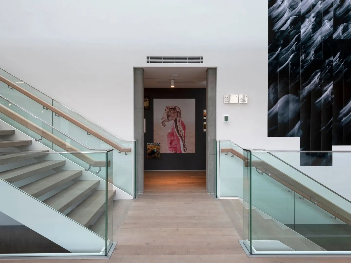 İstanbul'daki modern bir sanat galerisinin geniş giriş alanında, ortadaki geçide doğru uzanan şeffaf cam korkuluklu ve ahşap küpeşteli iki zarif merdiven dikkat çekerken, iç kısımda pembe giysili bir figürü betimleyen büyük bir tablo ve sağ duvarda dalgalı soyut formlardan oluşan etkileyici bir siyah beyaz sanat eseri sergileniyor.