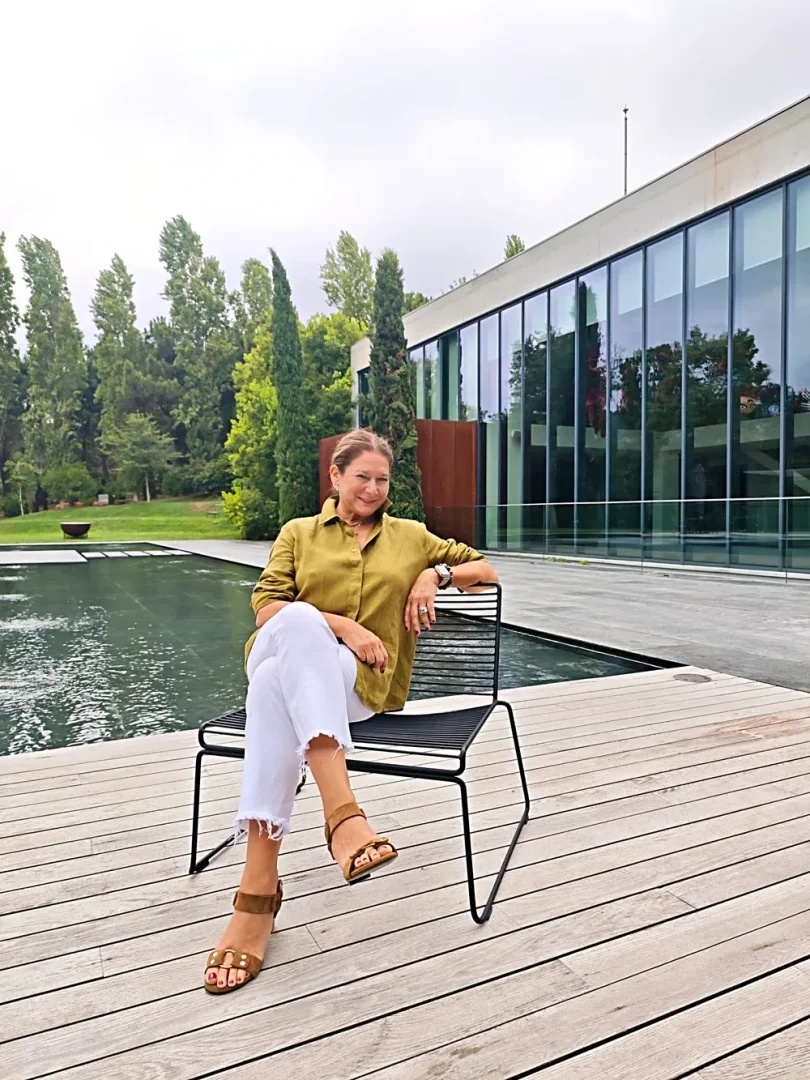 Hara İstanbul'daki modern cam cepheli bir binanın ve yemyeşil ağaçların önündeki ahşap terasta oturan Faika Ergüder, zeytin yeşili gömleği ve beyaz pantolonuyla şık bir sandalyede gülümseyerek objektife bakıyor.