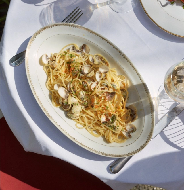 Beyaz masa örtülü bir masada, altın rengi kenarlı oval bir tabakta servis edilen, bol kabuklu istiridye, doğranmış maydanoz ve kırmızı pul biberle süslenmiş vongole spagetti, güneş ışığı altında keskin gölgelerle aydınlatılıyor; yanında çatal, bıçak ve şarap kadehi görülüyor.