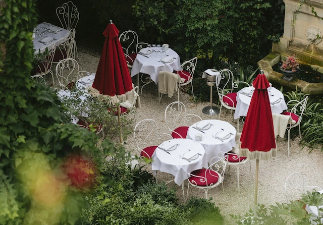 Hotel particulier'nin yemyeşil bahçesinde, beyaz masa örtülü yuvarlak masalar, kırmızı minderli ferforje sandalyeler ve kapalı kırmızı güneş şemsiyeleriyle donatılmış, arkada aslan başlı bir çeşmenin de göründüğü huzurlu ve şık bir açık hava restoranı alanı.