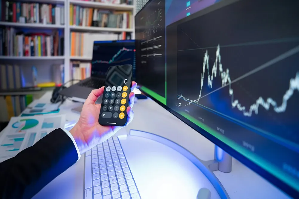 Bir kişinin hesap makinesi açık akıllı telefon tutan eli, karmaşık finansal grafikler ve verilerle dolu bir masaüstü monitörü ve dizüstü bilgisayarın önünde konumlanmış olup, bu görüntüsüyle döviz (FX) piyasalarındaki portföy yönetimi ve analiz süreçlerini net bir şekilde gözler önüne seriyor.