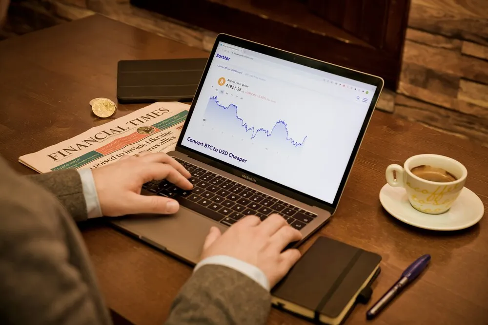 Bir kişi, Bitcoin/USD grafiği ve daha uygun FX dönüşümü bilgisi gösteren bir MacBook Pro dizüstü bilgisayarda aktif olarak tuşlara basarken, masanın üzerinde Financial Times gazetesi, altın rengi bir madeni para, sıcak bir fincan espresso ve not defteri gibi öğelerle finansal piyasalardaki portföy hareketlerini takip eden yoğun bir çalışma ortamı görülüyor.