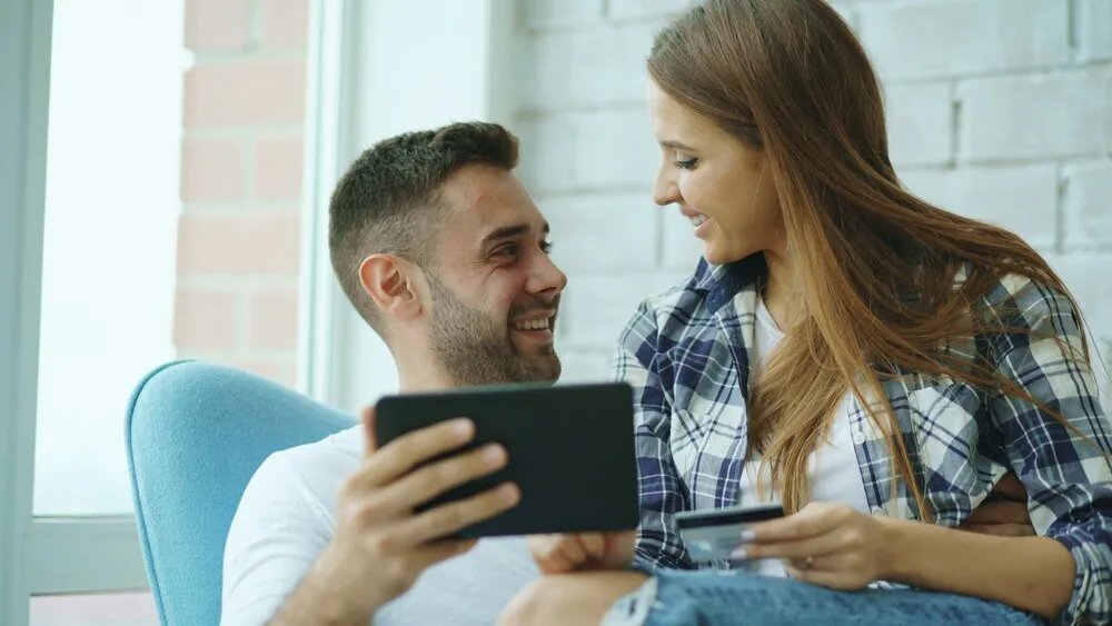 Genç bir çiftin, tablet ve kredi kartı ile finansal işlemlerini hallederken birbirlerine gülümseyerek baktığı, bir portföy yönetim kampanyasının avantajlarını düşündüren keyifli an.