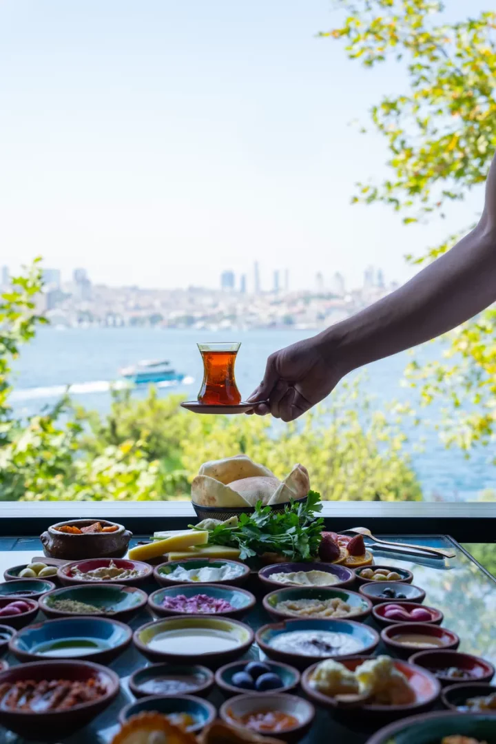 İstanbul Boğazı'nın eşsiz manzarasına karşı, cam bir masada, çeşitli peynirler, zeytinler, reçeller ve soslarla dolu sayısız küçük kaseden oluşan zengin bir serpme kahvaltı sunulurken, bir el zarifçe geleneksel bir çay bardağını uzatıyor ve arka planda İstanbul silüeti beliriyor.
