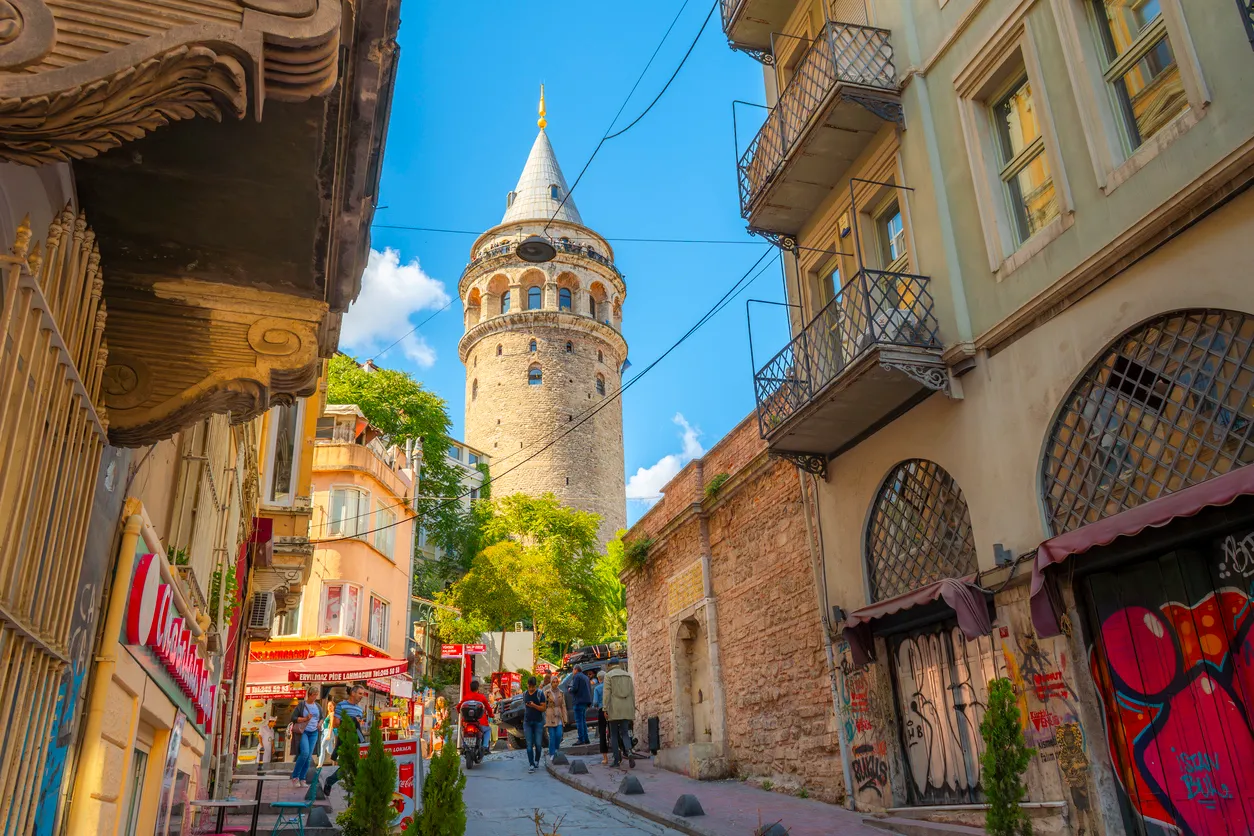 Mavi gökyüzü altında, dar ve tarihi bir yokuşta yürüyen insanların ortasında yükselen heybetli Galata Kulesi, İstanbul'un eşsiz yürüyüş rotalarından birinde, çevresindeki eski binalar ve canlı dükkanlarla birlikte şehrin kültürel zenginliğini ve hareketliliğini gözler önüne seriyor.