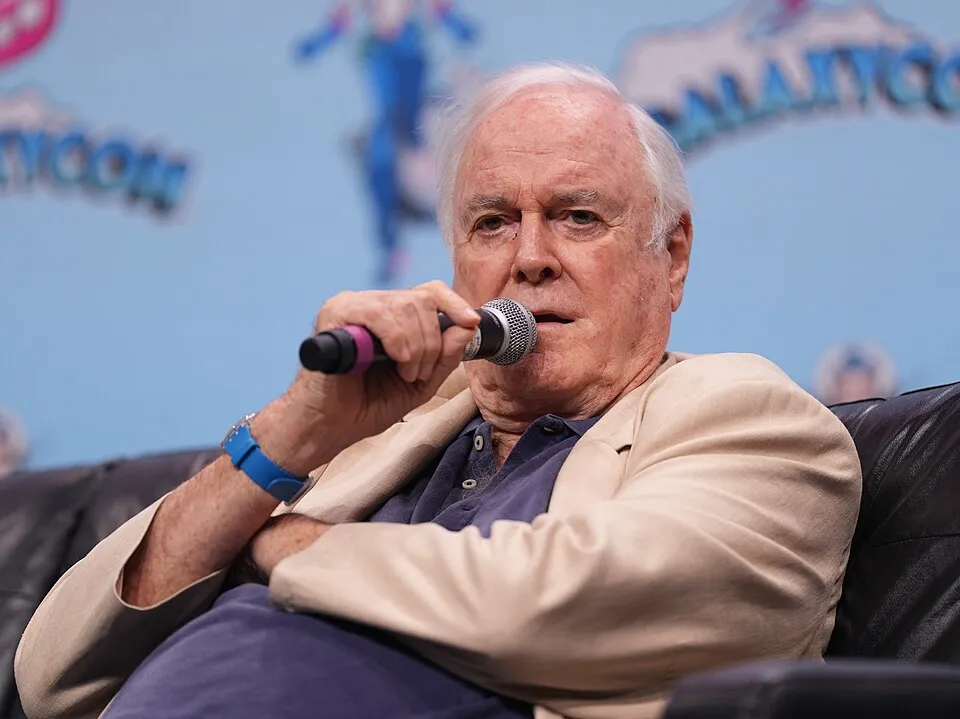Ortadan hafif açıyla çekilmiş bu karede, Britanya komedisinin efsane ismi John Cleese, açık renk ceketi ve lacivert polo yakalı tişörtüyle, elinde mikrofonu ağzına yakın tutmuş bir şekilde siyah bir koltukta otururken, arkasında bulanık bir etkinlik logosu görülüyor.