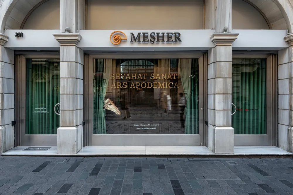Meşher'in mermer sütunlu ve kemerli modern girişi, cam vitrinde yeşil perdelerin arasından dışarı uzanan bir zürafa başı heykeli ve sokağın yansımalarıyla birlikte Seyahat Sanatı Ars Apodemica sergisinin adını belirgin bir şekilde sergiliyor.