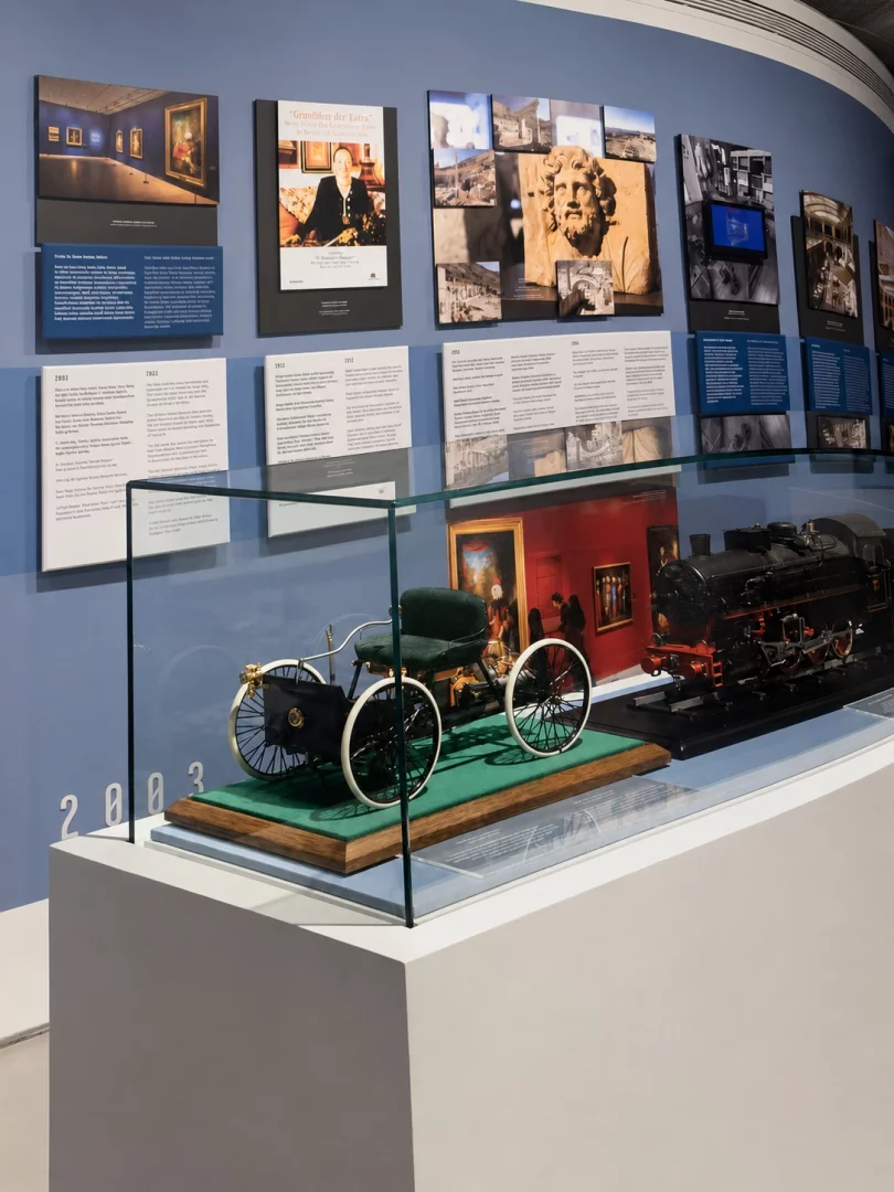 Yüzyılın İzleri: Koç Topluluğu ve Sanat sergisinden bir kesit sunan bu görselde, mavi duvara asılı kronolojik panolar ve çeşitli sanat eserleri ile tarihî görsellerin önünde, cam vitrinlerde eski bir otomobil modeli ve lokomotif maketi titizlikle sergileniyor.