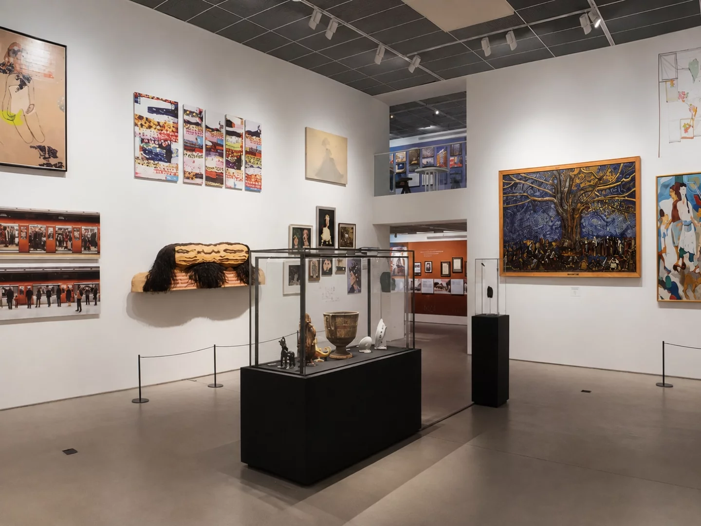 Beyaz duvarlı modern bir galeride, solda soyut resimler ve şehir hayatından fotoğraflar, ortada cam vitrinlerde heykeller ve antik eserler, sağda ise Aliye Berger'in kalabalık figürlü ve büyük ağaçlı ünlü tablosu dahil olmak üzere Koç Topluluğu ve Sanat sergisinden çeşitli dönemlere ait sanat eserleri dikkat çekiyor.