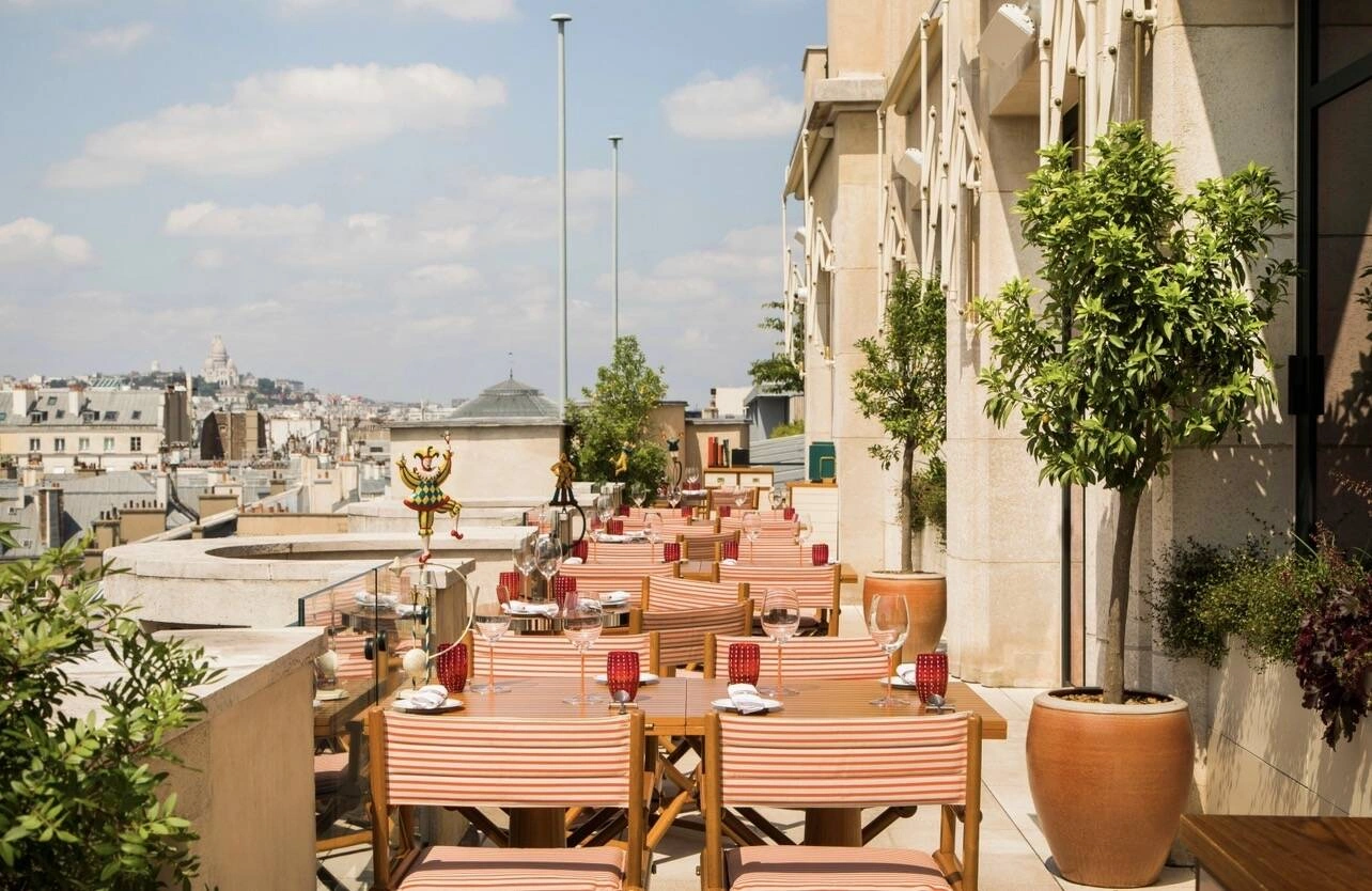 Langosteria Paris'in güneşli çatı terasında, kırmızı çizgili sandalyeler ve kadeh takımlarıyla zarifçe düzenlenmiş yemek masaları yer alıyor ve arka planda Sacré-Cœur Bazilikası'nın göründüğü Paris çatıları üzerinde büyüleyici bir panoramik manzara sunuluyor.