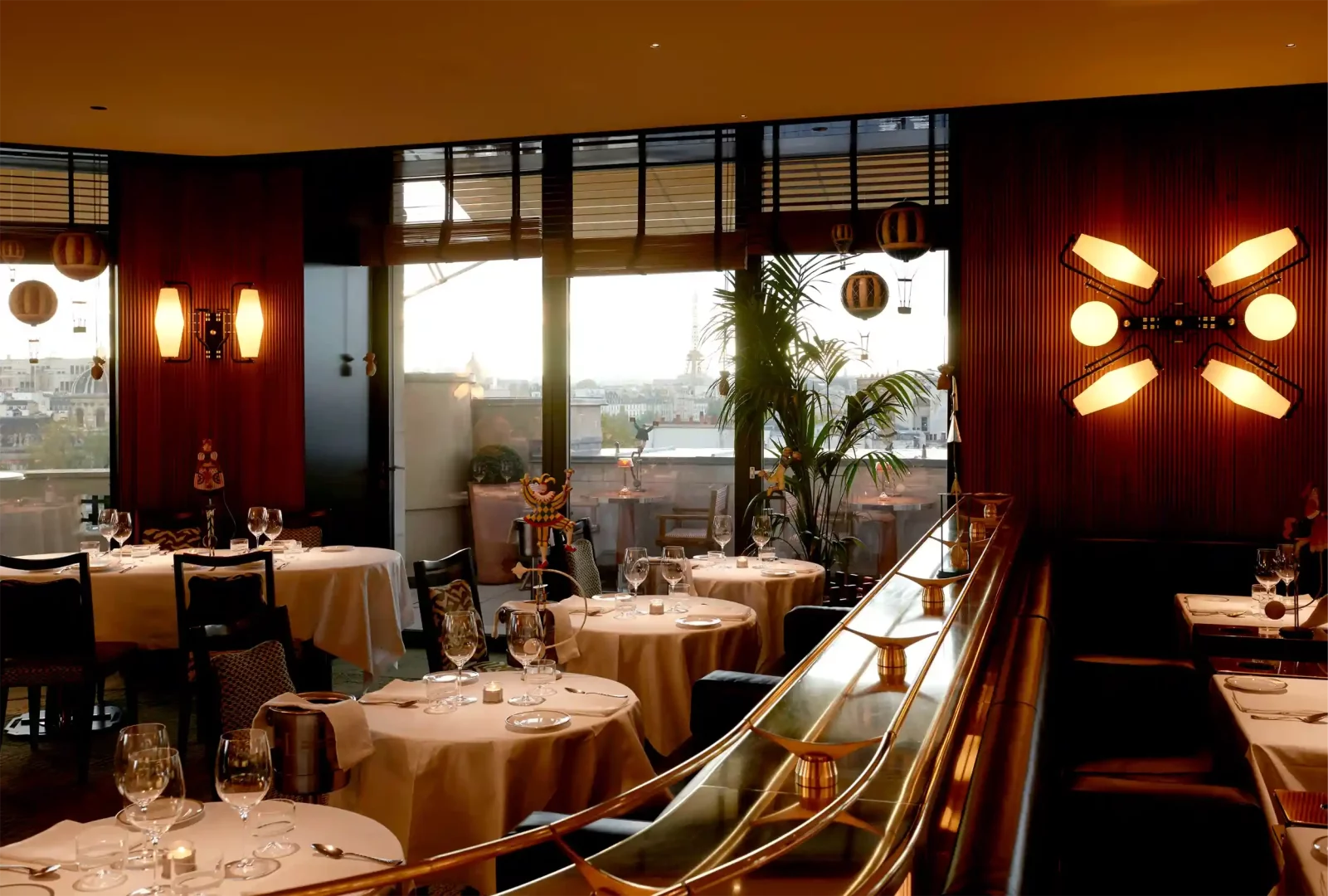 Langosteria Paris'in zarif yemek salonunda, pencereden görünen Paris şehir manzarası ve Eyfel Kulesi eşliğinde, beyaz örtülü masalar, şarap kadehleri, sıcak duvar aydınlatmaları, altın rengi detaylara sahip şık bir korkuluk ve tavandan sarkan dekoratif sıcak hava balonu figürleri yer alıyor.