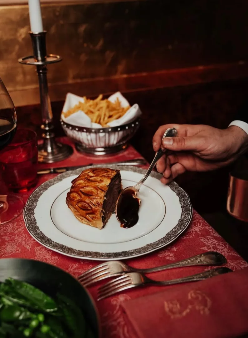 Loş ışıklı, romantik bir Paris restoranında, mum ışığında zarif bir date yemeği: bir el, dışı çıtır milföy hamurlu, içi et dolgulu yemeğe tabakta koyu renkli sosu dikkatlice eklerken, masada patates kızartması, kırmızı şarap ve yeşil sebzeler bu Fransız klasiğini tamamlıyor.