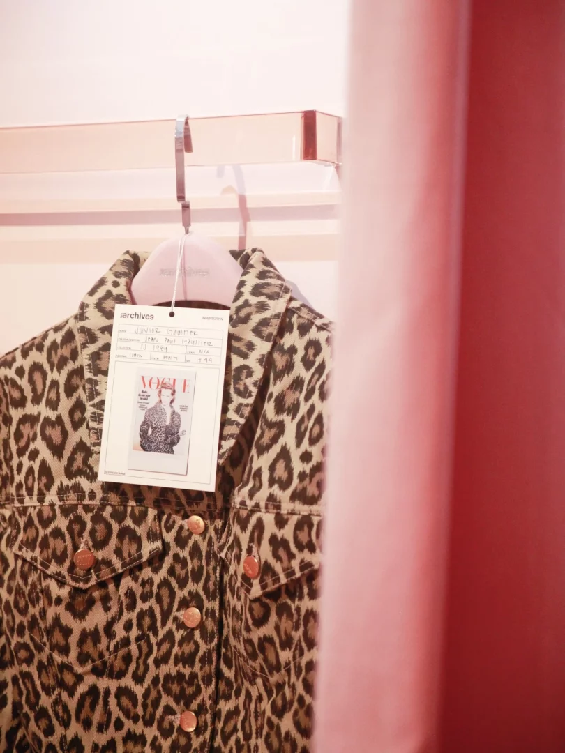 Pembe bir askılıkta asılı duran, bakır renkli düğmeleri olan leopar desenli bir ceketin üzerinde, Jean Paul Gaultier'in 1989 Junior Gaultier koleksiyonundan bir parça olduğunu belirten, üst kısmında arşivler yazan ve Vogue dergisinden bir görsel içeren beyaz bir etiket görülüyor.