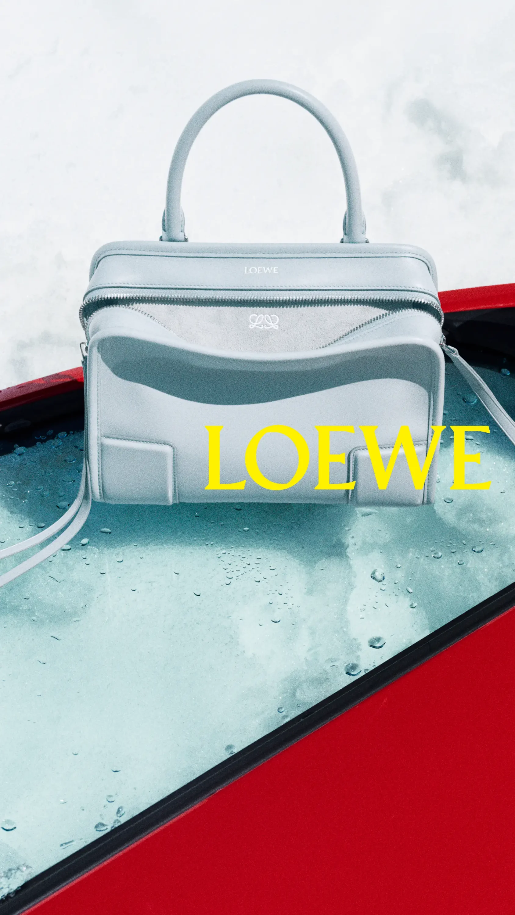 Açık buz mavisi renkte, üstten kulplu ve omuz askılı, Loewe logosu taşıyan zarif bir çanta, yağmur damlalarıyla ıslanmış cam gibi görünen bir yüzey üzerinde dururken, bu fotoğraf eylül ayında öne çıkan markaların yeni tasarımlarından birini yansıtıyor.