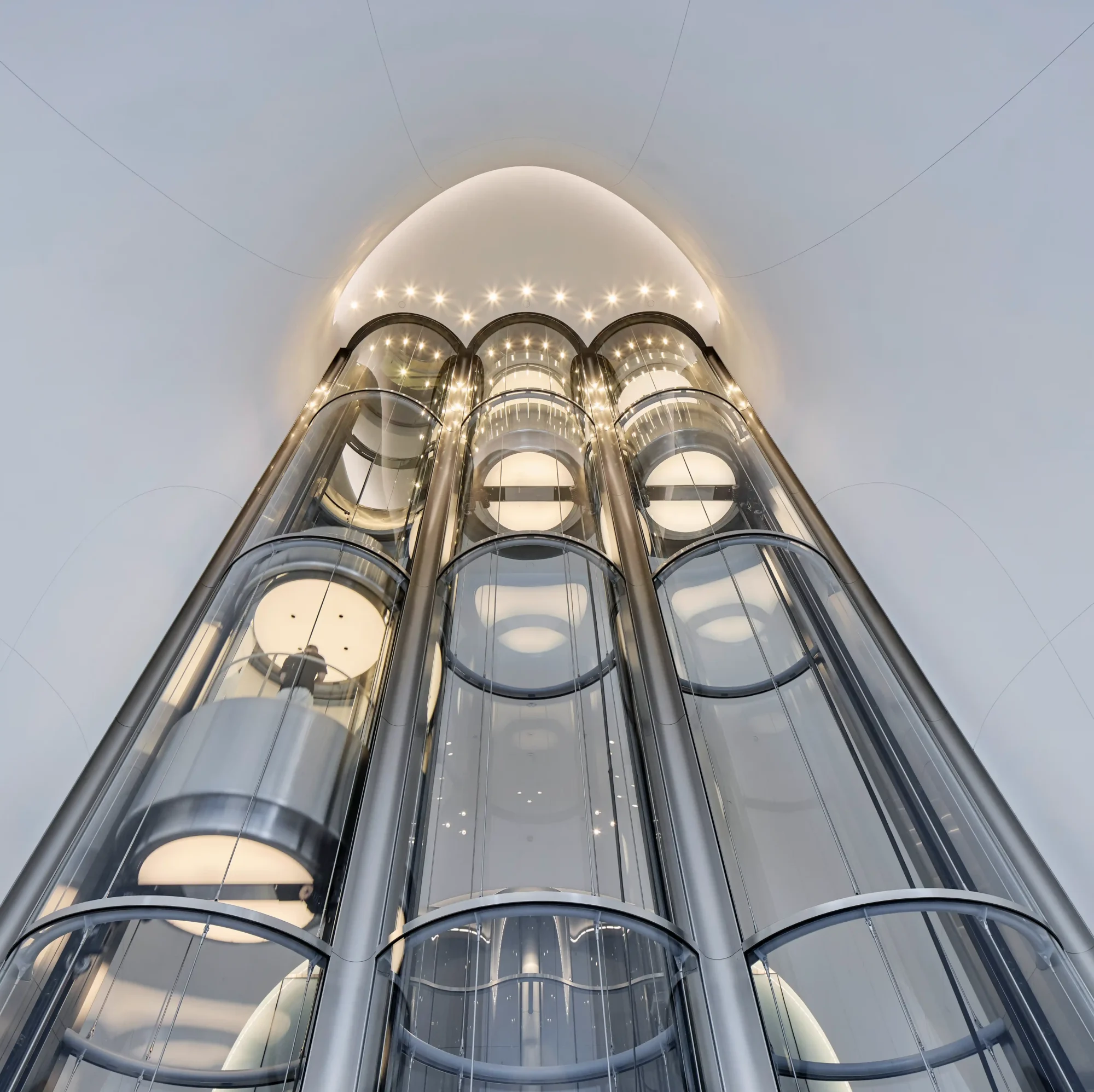 Lucas Museum of Narrative Art'ın kuzey lobisinde, ziyaretçileri üst katlardaki hikaye galerilerine taşıyan ve bu yükselişi mimari bir deneyime dönüştüren üç silindirik cam asansör, parlak bir tavan altında yukarı doğru süzülürken içindeki yolcuların siluetlerini gözler önüne seriyor.
