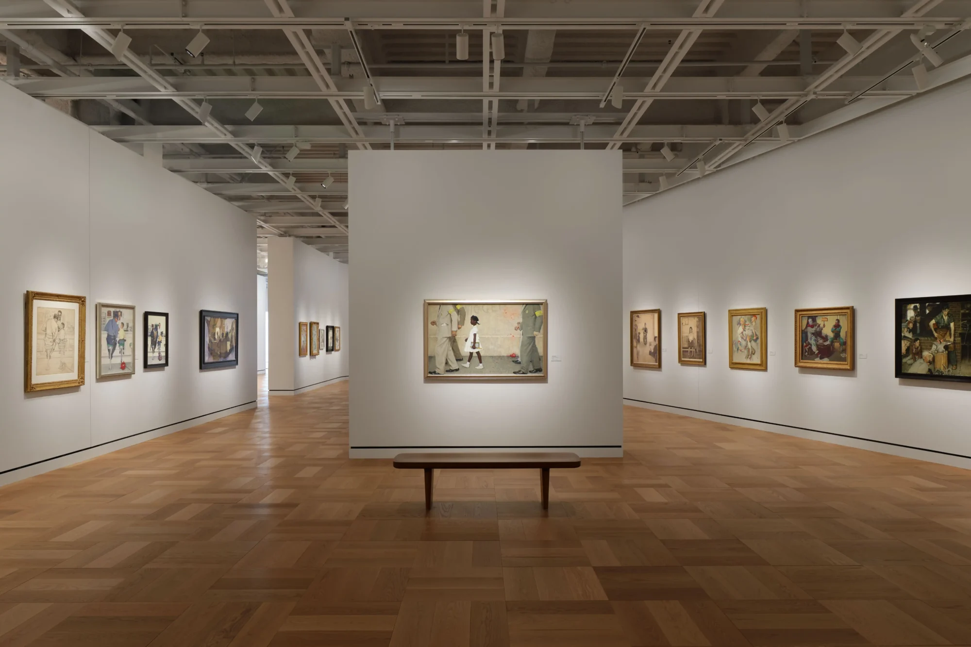 Modern ve geniş bir sanat galerisinin parke zeminli aydınlık salonunda, beyaz duvarlarda sergilenen farklı boyutlardaki çerçeveli tablolar, Lucas Anlatı Sanatı Müzesi'nin görsel hikaye anlatıcılığına odaklanan zengin koleksiyonunu yansıtıyor; merkezdeki duvarda tarihi bir olayı betimleyen büyük bir tablonun önünde uzun, ahşap bir bank yer alıyor.