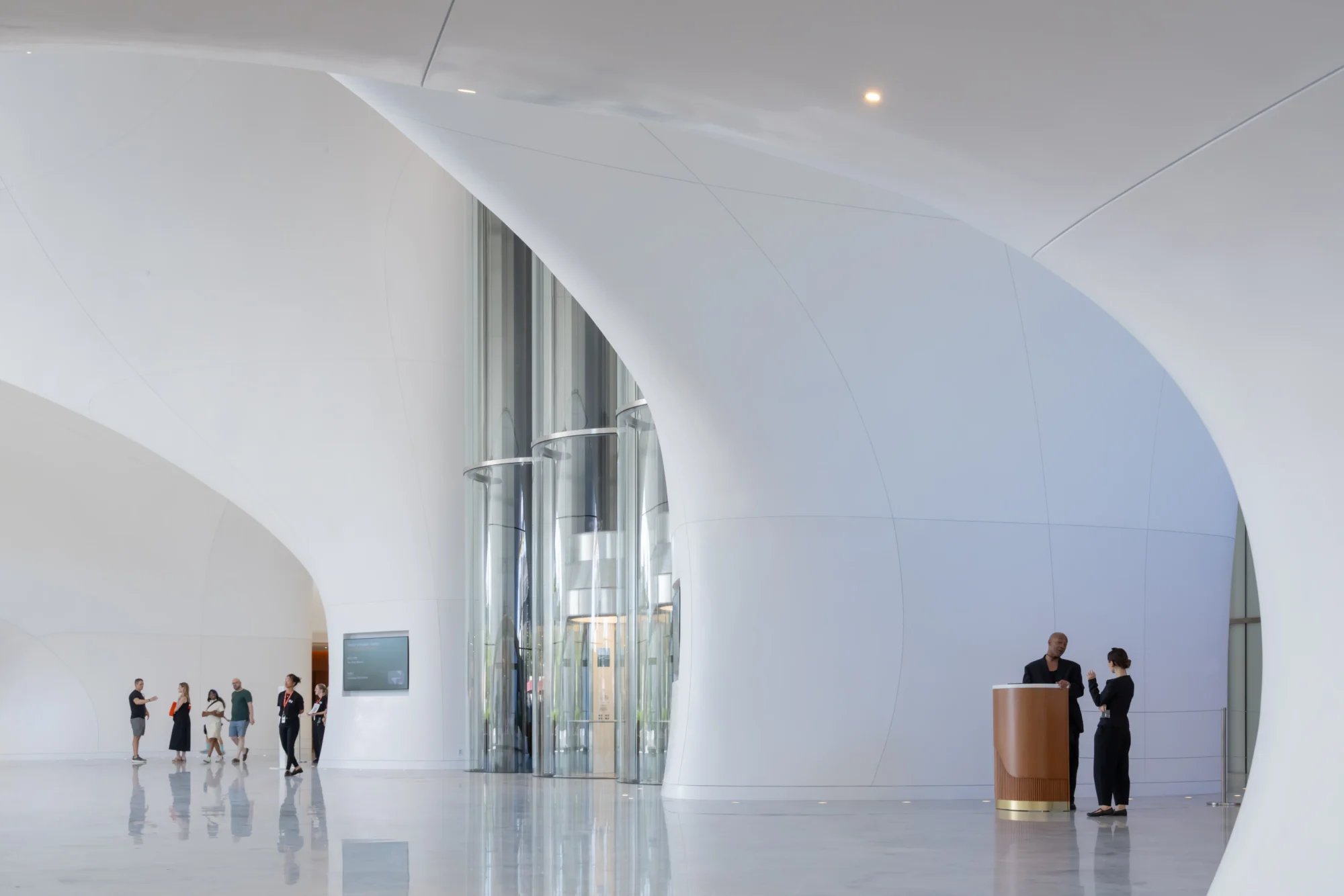 George Lucas'ın kurduğu Lucas Museum of Narrative Art'ın Ma Yansong tasarımı mimarisini yansıtan, bulut hafifliğindeki akıcı kavisli beyaz duvarlar ve yansıtıcı zeminle çevrili bu geniş ve aydınlık ana lobide, ziyaretçileri görsel hikaye anlatıcılığının zengin dünyalarına taşıyan üç silindirik cam asansörler ve ahşap bir danışma masası görülüyor.
