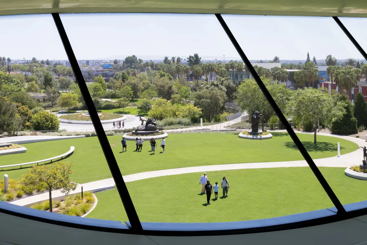 Lucas Museum of Narrative Art'ın modern mimarisine ait geniş, kavisli penceresinden görünen Los Angeles'taki yemyeşil parkta, George Lucas'ın hikayelerle dolu dünyasını temsil eden heykeller arasında gezinen ziyaretçiler ve uzaktaki şehir silueti yer alıyor.