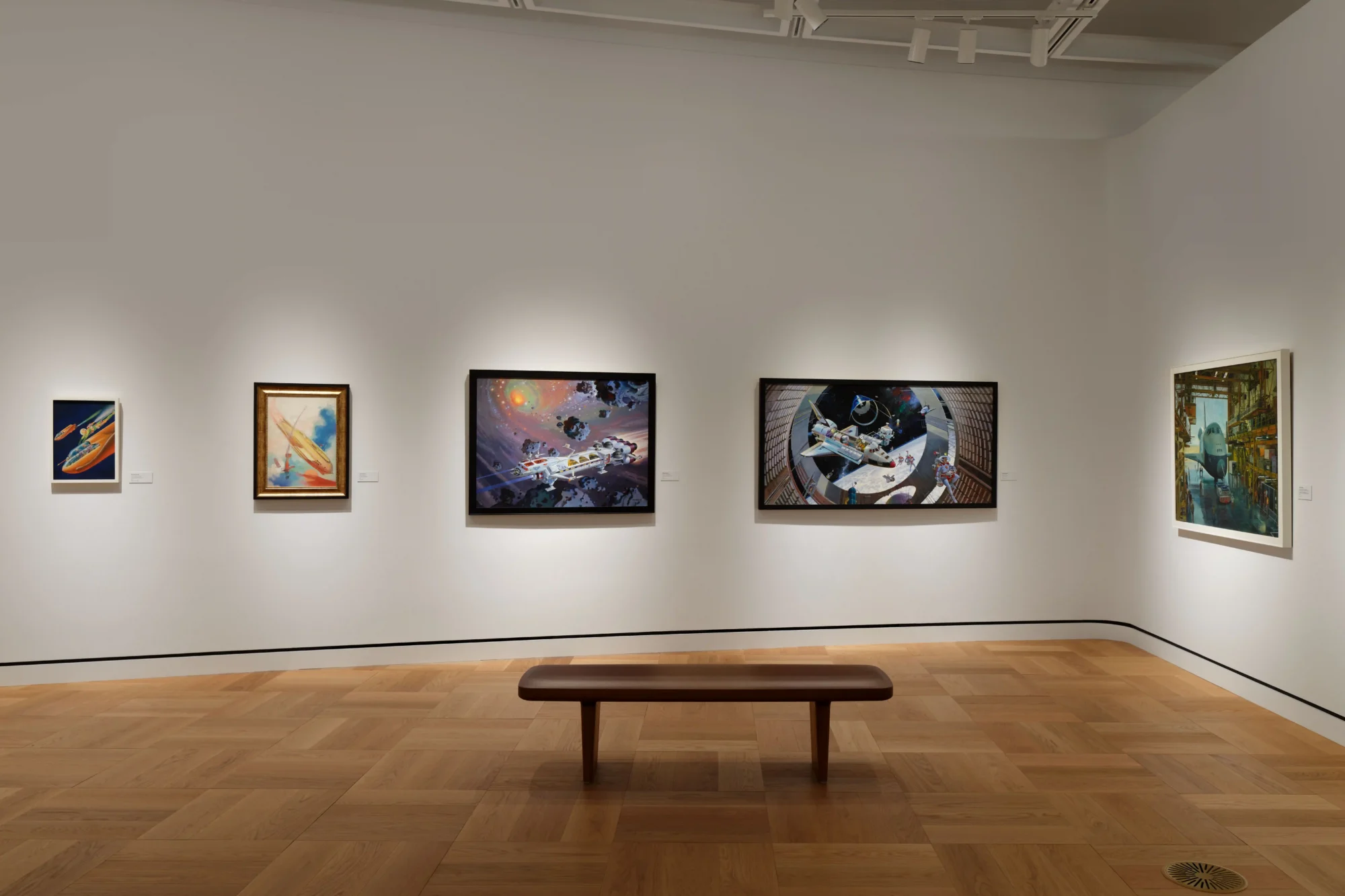 Beyaz duvarlı bir sanat galerisinde, ortada ahşap bir bankın önünde asılı duran, uzay gemilerini ve hayali dünyaları betimleyen beş farklı boyut ve çerçevedeki görsel anlatı eserleri, George Lucas'ın biriktirdiği eşsiz koleksiyondan parçalar sunuyor.