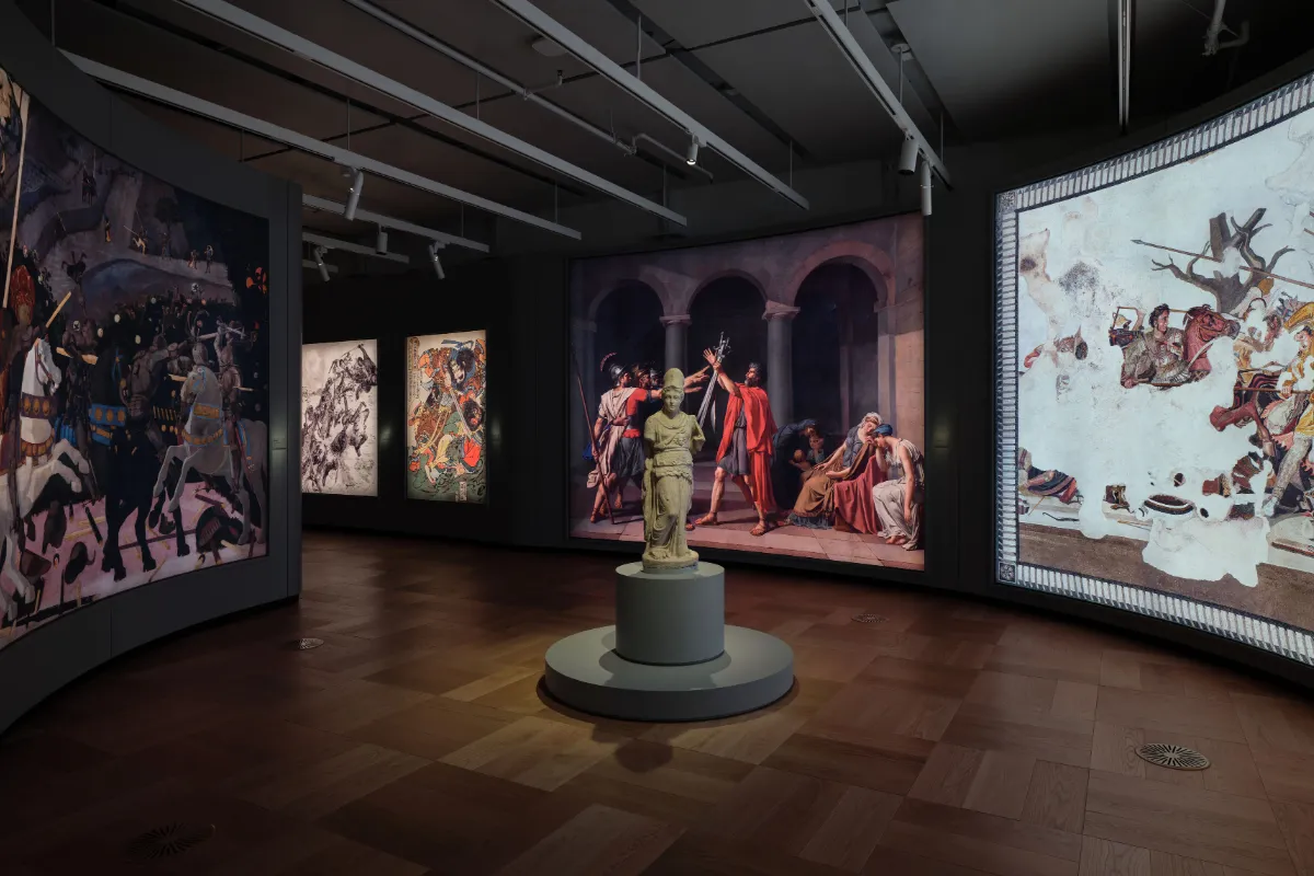 Lucas Museum of Narrative Art'ın geniş bir galerisinde, zengin anlatı sanatı koleksiyonundan çarpıcı eserler sergileniyor: Ortada mermer bir heykelin arkasında dramatik bir antik dönem tablosu, solda atlı savaşçıların olduğu bir resim ve Japon çizimlerine benzeyen eserler, sağda ise antik bir mozaik gibi duran büyük bir savaş sahnesi bulunuyor, hepsi farklı hikayeleri görselleştiren sanat eserlerinin çeşitliliğini gözler önüne seriyor.
