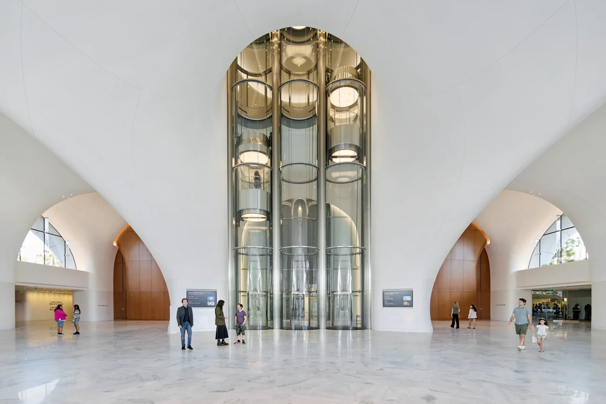 Lucas Museum of Narrative Art'ın bulutları andıran beyaz, geniş ve ferah lobisinde, dördüncü kattaki anlatı sanatı galerilerine ziyaretçileri taşıyan üç parlak silindirik cam asansör ve çevresinde dolaşan insanlar yer alıyor.