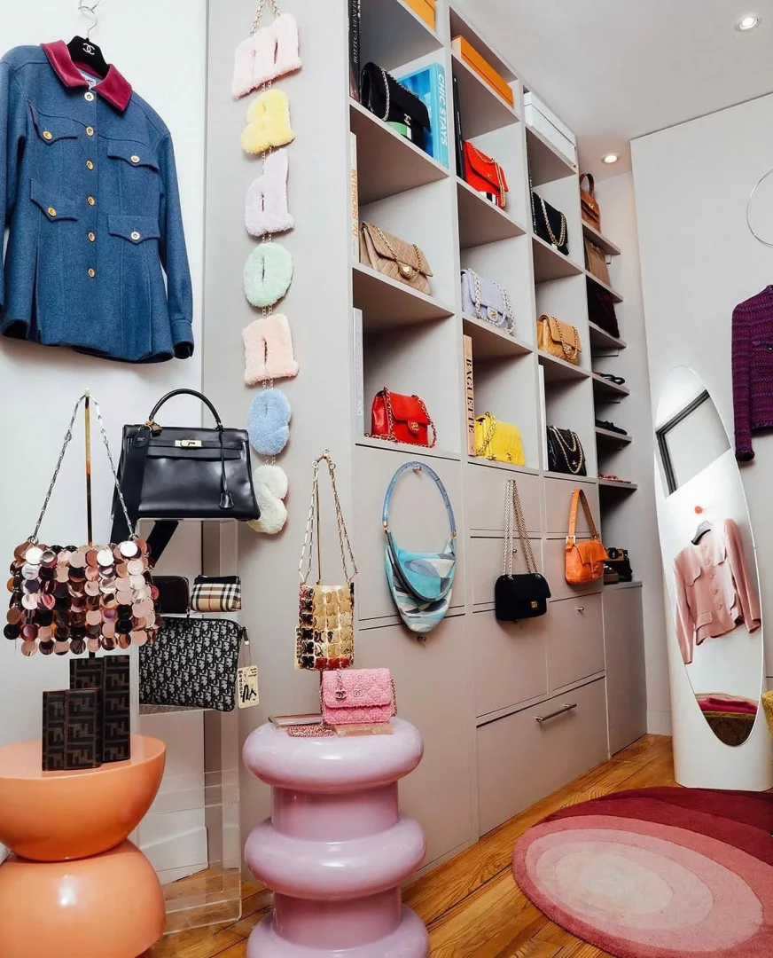 Canlı ve titizlikle düzenlenmiş bir lüks giyinme odasında, raflarda sergilenen, duvarlarda asılı ve şık tabureler üzerinde duran çok sayıda tasarımcı çanta, MADONES yazan renkli peluş harf çantalar ve pembe tonlarında bir halı göze çarpıyor.