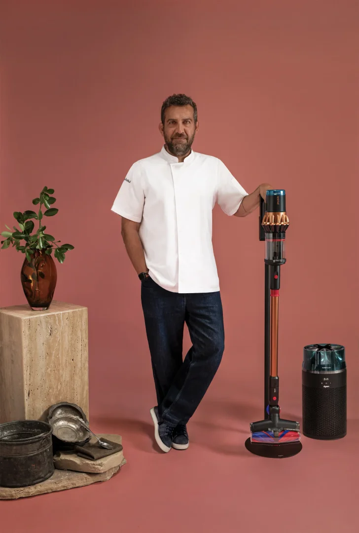 Beyaz şef ceketli ve kot pantolonlu Maksut Aşkar, gülümseyerek, sağ eliyle modern bir Dyson dikey süpürgeye yaslanmış; yanında bir Dyson hava temizleyici, arkasında ise geleneksel mutfak eşyaları ve bitkili bir vazo, hijyen ve çağdaş yaşam dengesini yansıtan bir kompozisyon oluşturuyor.