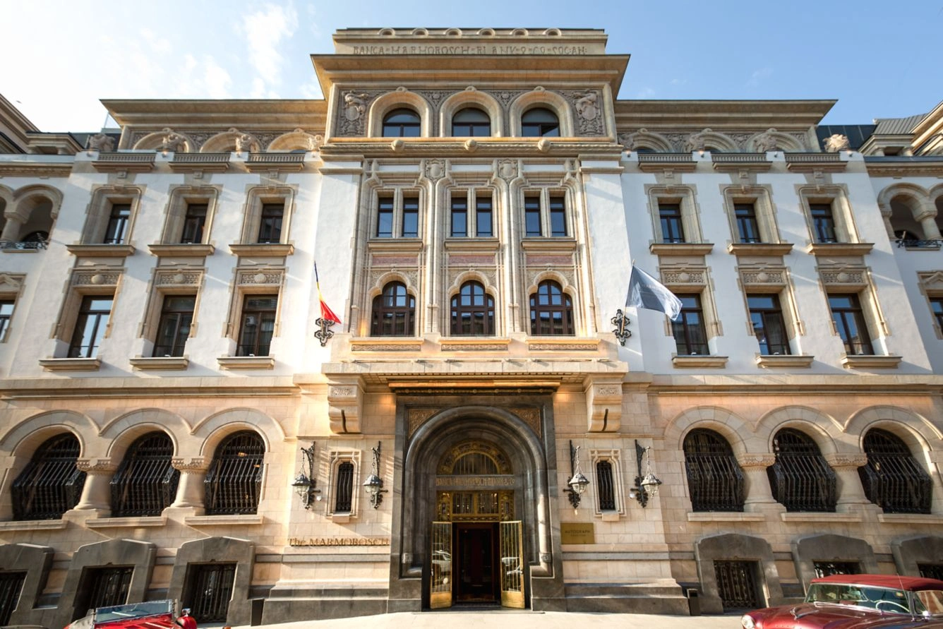 Güneşli bir günde çekilen bu geniş açılı görselde, eski bir banka olan Marmorosch binasının Art Nouveau esintili, taş ve beyaz sıvalı, zengin süslemeli tarihi cephesi önünde, solunda kırmızı bir üstü açık araba ve sağında koyu renk bir klasik otomobil ile iki bayrak dalgalanıyor.