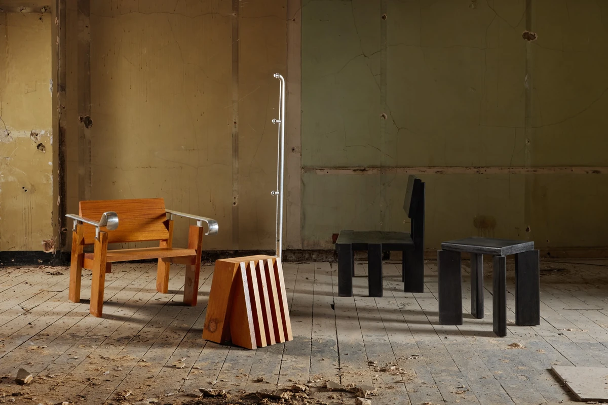 London Design Festival 2026'daki Material Matters sergisinde yer alan, yıpranmış duvarları ve tozlu zemini olan endüstriyel bir ortamda, ahşap ve metalden yapılmış modern tasarımlı bir koltuk, çizgili ahşap bir blok, metal bir askılık, koyu renk ahşap bir sandalye ve küçük bir tabureden oluşan Prima Materia enstalasyonu.