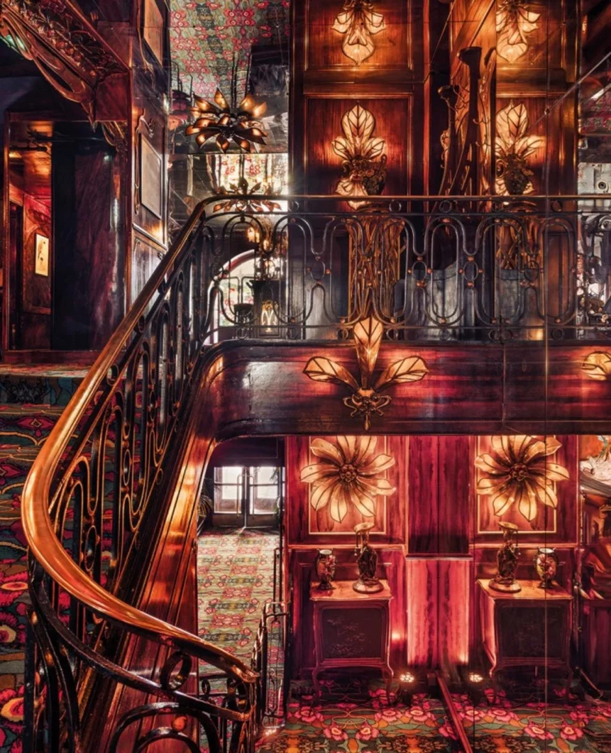 Art Nouveau tarzında dekore edilmiş, koyu ahşap paneller, altın rengi ferforje detaylar ve çiçek desenli halılarla zenginleştirilmiş gösterişli bir iç mekanda, parlak pirinç tırabzanlı ve işlemeli metal korkuluklu, yukarı doğru kıvrılan büyük bir merdiven, duvarlarda yaprak motifli aydınlatmalar ve aynalı yüzeyler çarpıcı bir atmosfer yaratıyor.