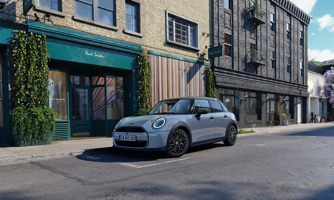 Açık gri Mini Cooper Paul Smith Edition otomobil, yeşil renkli Paul Smith mağazasının önündeki şehir caddesinde park halinde duruyor; arabanın tavanında ve ayna kapaklarında zarif yeşil detaylar göze çarpıyor.