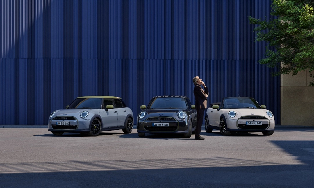 Modern mavi panelli bir binanın önünde, özel sarımsı yeşil detaylara sahip gri bir 5 kapılı, siyah bir coupe ve krem rengi bir cabrio Mini Cooper Paul Smith Edition otomobilleri park edilmiş; şık takım elbiseli bir adam bu farklı model araçları düşünceli bir ifadeyle inceliyor.
