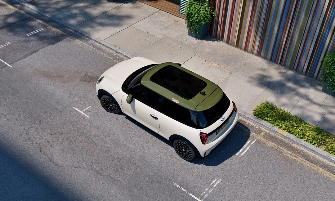 Kuşbakışı çekilmiş bu fotoğrafta, özel Paul Smith Edition tasarımına sahip, zeytin yeşili tavanlı ve geniş güneşlikli beyaz Mini Cooper, asfalt bir yolda park edilmiş olarak görülüyor; aracın sağında kaldırım, yemyeşil çalılar ve dikey renkli şeritleri olan modern bir bina duvarı yer alıyor.